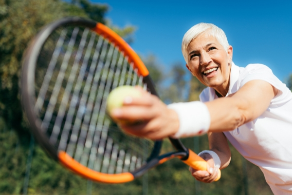 An elderly woman holding a tennis racquet and tennis in a serving position.